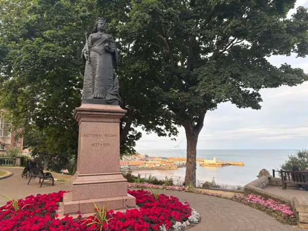 Statue of Queen Victoria on a granite pedestal surrounded by flower beds, with Scarborough’s seafront and townscape visible in the background.